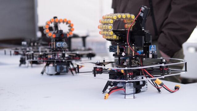 Quadcopter drone prototypes with clear protective frames and exposed electronics on white table, one unit in sharp foreground and blurred workshop background