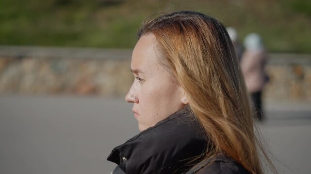 Woman with long hair and black jacket is shown in profile looking away from camera. background features outdoor setting with greenery and stone wall elements suggesting daylight conditions