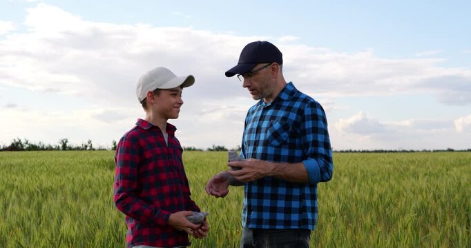 Farmer father gives work gloves to son in green wheat field before farm work. Scene symbolizes passing agricultural knowledge to next generation, family tradition, rural heritage and shared work.