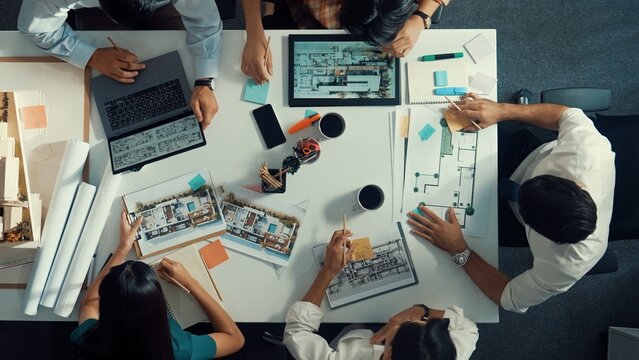 Top view of smart architect engineer writing at blueprint while sitting at meeting table with house model and laptop. Skilled manager working and taking about about building structure. Alimentation.