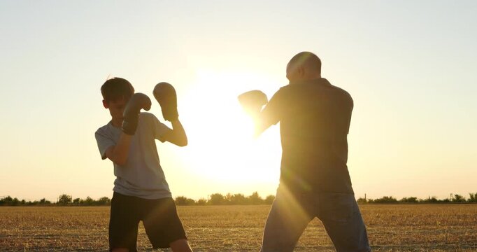 Dad and son practice boxing together in open field at sunset. Father and child train with focus, fitness and teamwork. Outdoor family workout shows playful sparring, discipline and active lifestyle.