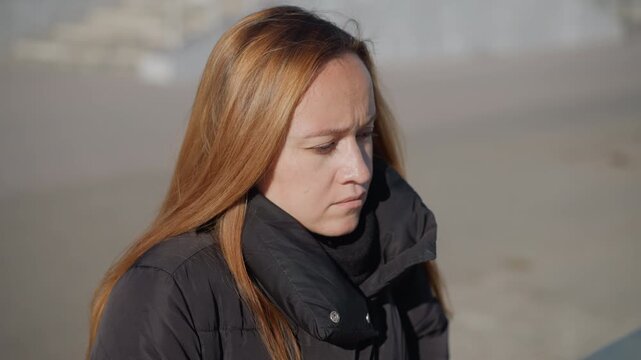 Woman with long light brown hair wears black puffer jacket and stands outdoors in soft daylight. Her expression is neutral to slightly introspective as woman looks off camera suggesting
