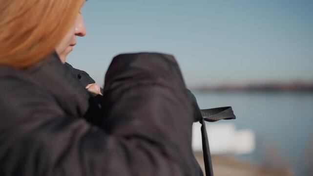 Woman outdoors near water handling black bag and retrieving sunglasses. Woman wears dark clothing with long hair visible from side as woman interacts with her personal items against outdoor