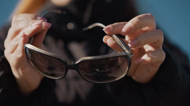 Woman holds and examines pair of sunglasses displaying them prominently in close up. Her hands with manicured nails are visible as woman adjusts glasses revealing details like dark lenses