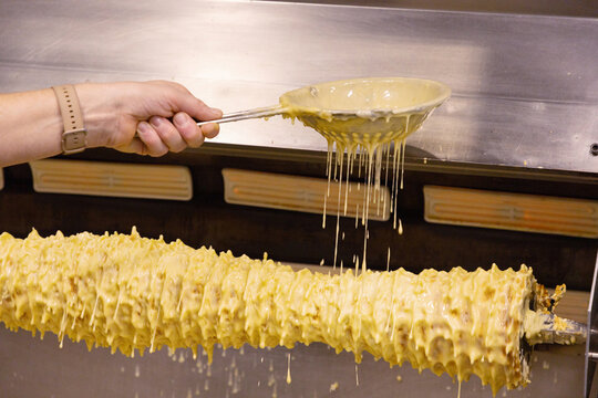 Hand pouring sauce over rotating meat skewer in commercial kitchen setting