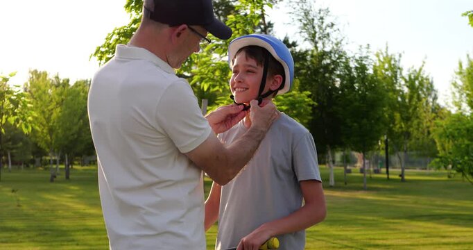 Father helps son adjust bicycle helmet in sunny park. They prepare for outdoor activity with care, trust, and family support. Warm summer scene promotes parenting, safety, and healthy lifestyle