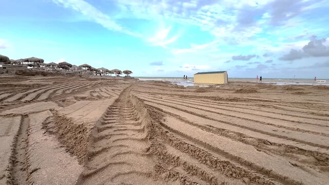 Deep tire tracks on the wet sand of Vama Veche beach at low tide with thatched umbrellas, a flooded container, and soft daylight over the Black Sea coast. 4K video.