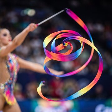 Rhythmic gymnast swirling colorful rainbow ribbon during indoor competition performance.