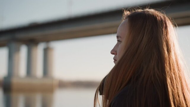Woman with long brown hair stands outdoors near body of water and overpass looking into distance. scene features soft natural light reflecting on her face against calm surroundings