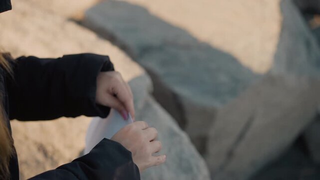 Woman in black jacket examines and unfolds white paper against outdoor rocky background. Her hands manipulate object with natural light illuminating her skin and surroundings. scene focuses