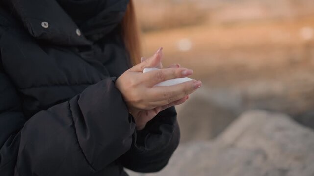 Woman in black jacket holds and manipulates folded white paper against outdoor background with soft natural light. Her hands are primary focus showing interaction details like grasping
