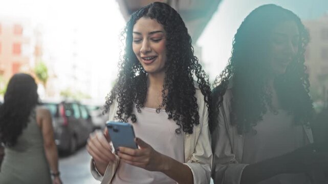 Young arab woman holding smartphone and smiling, fist raised in a casual street scene reflected in storefront glass; joy connection.