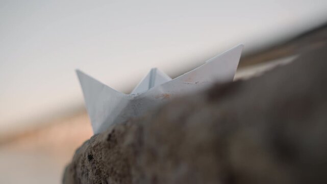 White paper boat rests on rough stone surface with soft natural light. background is blurred emphasizing texture of both folded material and rock. scene maintains consistent focus throughout