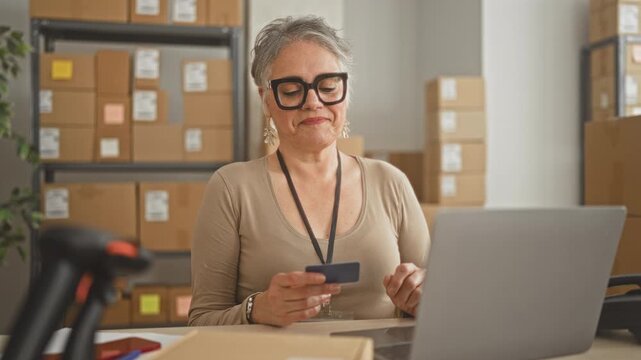 Woman holding creditcard and smiling while typing on laptop amid stacked boxes and barcode scanner in building; small business confidence.