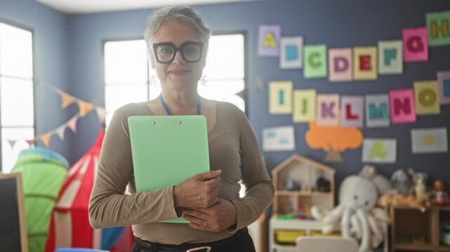 Woman teacher with short hair and glasses holding a mint clipboard, smiling and clasping it in a colorful classroom decorated with alphabet cards and toys; nurturing care education.