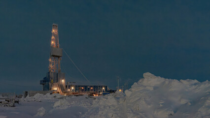 Illuminated oil and gas drilling rig operating in the harsh snowy Arctic landscape during winter polar night. Industrial energy extraction and exploration concept. © vladend