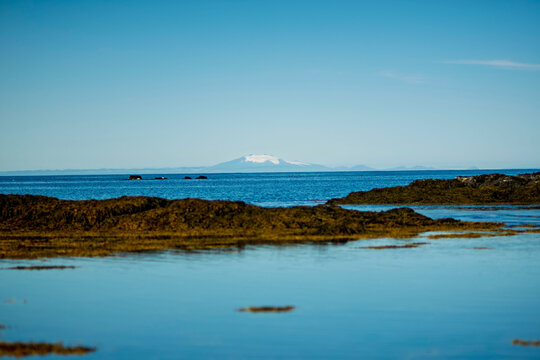 Distant snow capped glacier volcano over blue ocean horizon and tide pool in Akranes Iceland