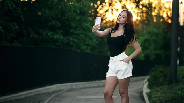 Beautiful attractive young woman taking selfie on smartphone in park at sunset. Pretty brunette female posing and holding mobile phone outdoors with bright lens flare background