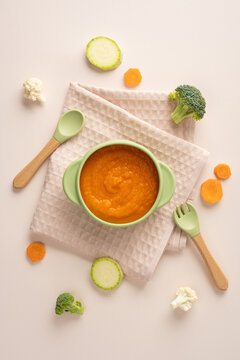 Baby first feeding with healthy carrot puree in a green bowl with baby spoon and vegetable on vertical pastel table