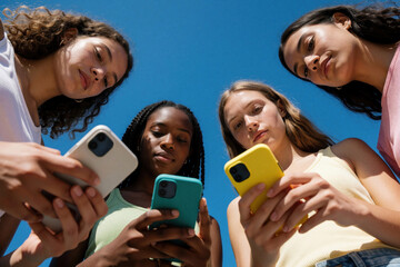 Obraz premium Four young women hold smartphones looking down at screens. Friends connect via mobile phones under clear blue sky. They browse content, share info, enjoying time together.
