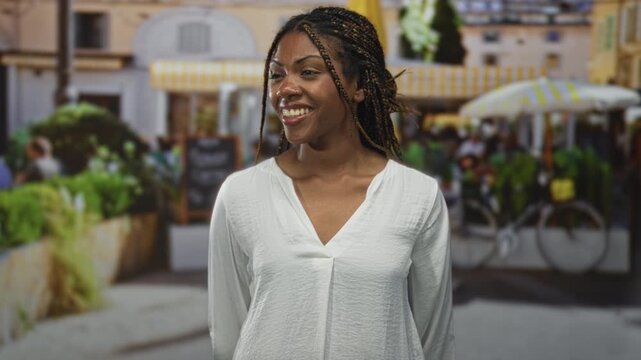 African american woman smiling with braided hair and nosepiercing, wearing a white blouse, standing in a street cafe near bicycles and planters; joy confidence warmth.