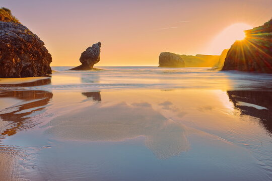 Sunstars illuminate 'El Pic&oacute;n' limestone pinnacle on a vibrant fiery dawn, reflected on Playa de Buelna Beach at low tide. Llanes-Asturias-Spain-081