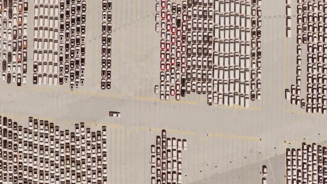 Top-down aerial view of a huge parking lot filled with brand new cars ready for export, showing the scale of the automotive industry's logistics and global trade network