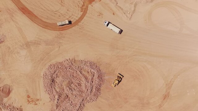 Top down aerial perspective showing a yellow bulldozer pushing gravel while heavy dump trucks maneuver around the vast, arid landscape of an open-pit mine during a sunny day