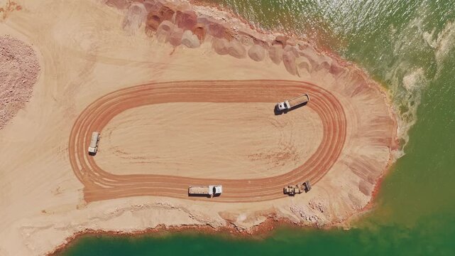 Top-down aerial view of heavy machinery operating in a large open-pit mine, with dump trucks transporting materials along a dirt track next to a green body of water in an industrial area