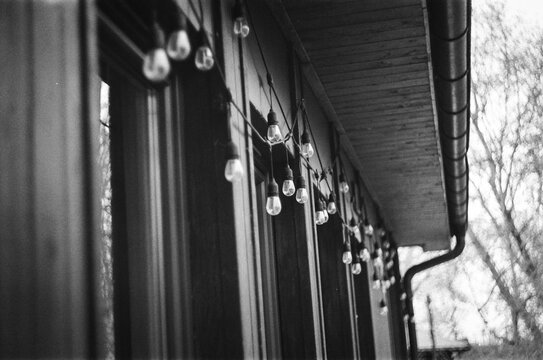 Black and white photo of string lights and light bulbs hanging under the eaves of a wooden house