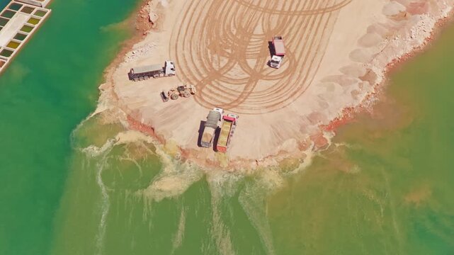 High angle view of a tipper truck working at a construction site, backing up to the edge of a river or lake and unloading sand to create new land for industrial development