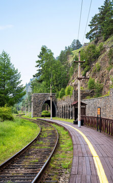 Vertical photo of a curving railway track leading into a stone tunnel at Lake Baikal, Siberia. Woden passenger platform next to a rocky cliff. Summer nature landscape with old railroad