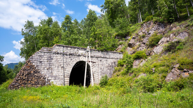 Old stone tunnel entrance built into a rocky hillside at Lake Baikal, Russia. Historic arched railway passage surrounded by green summer vegetation and trees. Abandoned infrastructure in nature