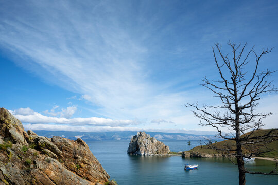 Landscape view of a rocky island, Shamanka Rock on Lake Baikal in Siberia. Bare tree and rugged cliff in the foreground with a small boat on the water. Blue sky background