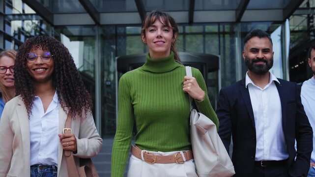 A group of cheerful young professionals walks forward outside a contemporary corporate venue. They are smiling and looking at the camera during a bright daytime work break