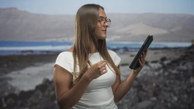 Woman holding tablet with hand on chest reading from screen while wearing glasses in studio coastal set; quiet contemplation.