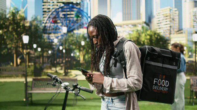Impatient bicycle courier holds a smartphone and thermal delivery backpack in an urban park, anxiously waiting for a new order request on a mobile app during app-based gig delivery work.