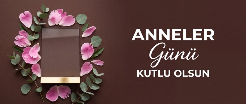 Anneler Gunu Kutlu Olsun Turkish Mother's Day banner with elegant glass frame, gold stand, pink flower petals and green eucalyptus leaves on dark brown background for elegant holiday celebration mocku
