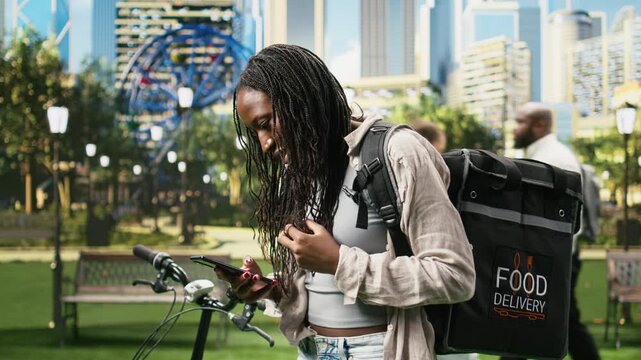 Anxious woman courier fidgets while waiting for a new order request, repeatedly checking a mobile delivery app beside her bike and thermal bag in a park during gig-economy food delivery work.