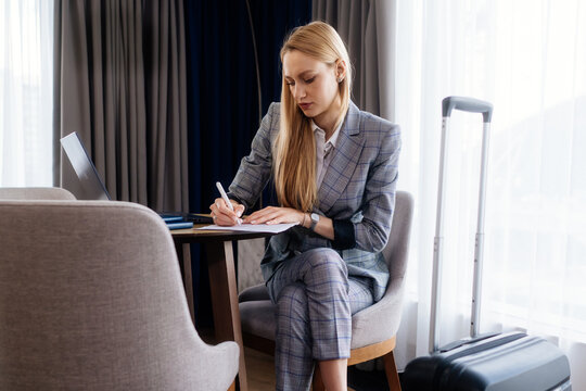 Businesswoman writing documents beside laptop and suitcase for work. The table becomes a temporary workspace during business travel.