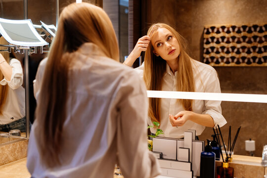 Woman combing long blonde hair in bathroom mirror for self-care. Vanity lighting frames a detailed self-care activity.