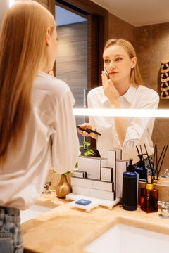Woman removing excess facial oil with mattifying blotting paper. She watches her reflection while refreshing her complexion at the bathroom vanity.