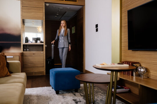 Businesswoman in formal suit entering hotel room with suitcase. She crosses the doorway at the start of a corporate trip.