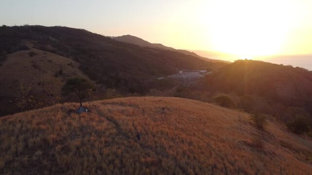 Cinematic aerial footage of golden grass-covered hills during sunset, with layered mountain ridges and warm sunlight creating a peaceful natural landscape.