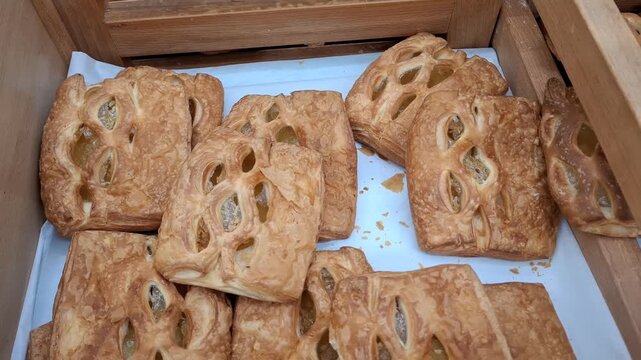 Golden puff pastries lie in a wooden crate indoors. Fresh bakery goods, sweet danish pastry, bakery store display.