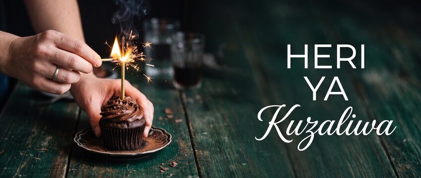 Chocolate cupcake birthday sparkler candle being lit by hands with Swahili Heri ya Kuzaliwa text on rustic green wooden table background, moody dark celebration card graphic banner copy space