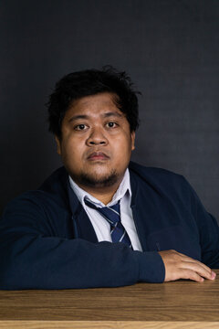 A startled young Asian male professional sits stiffly at a smooth wooden desk, displaying a wide eyed, stunned facial expression against a seamless black studio background. Dressed