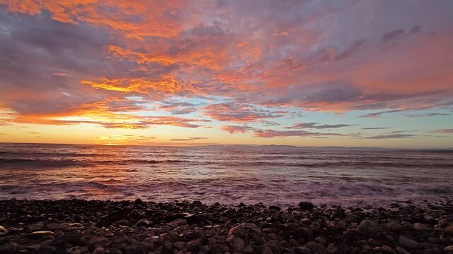 Fiery clouds paint the sky as waves roll onto a pebble beach during a breathtaking sunset over the sea