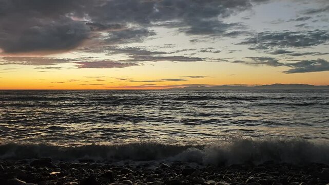 The sun dips below the horizon casting a warm glow on the sea as waves gently roll onto a pebble beach
