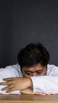 A young Asian male office employee rests the head completely face down on crossed arms over a smooth wooden desk against a solid dark gray wall. The exhausted corporate professional